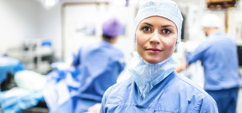 En sjuksköterska står i en operationssal. I bakgrunden pågår en operation.